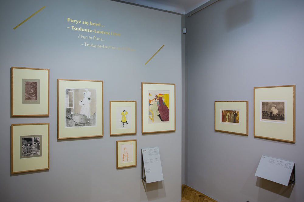 Narożnik galerii z jasnoszarymi ścianami, na których w drewnianych ramach z kremowymi passe-partout rozmieszczono dziewięć grafik — kolorowych i czarno-białych —, dwa stojące stoliki z opisami ekspozycji na podłodze oraz na ścianie widoczny żółty napis Paryż się bawi... — Toulouse-Lautrec i inni