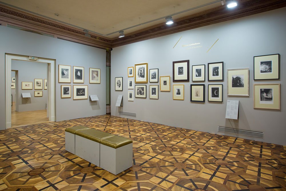 Wnętrze galerii z jasnymi ścianami prezentujące liczne oprawione czarno-białe fotografie i grafiki w różnych ramach rozmieszczone na dwóch ścianach, ozdobna mozaikowa drewniana podłoga, prostokątna ławka z zieloną poduszką na środku, punktowe oświetlenie sufitowe i otwarte przejście do sąsiedniej sali.