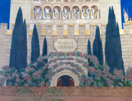 Fragment murów i baszty z jasnego kamienia z arkadowym balkonem z sześcioma figurami, kamienną tablicą z napisem Turris Eburnea nad półokrągłym wejściem obrośniętym pnączami z różowymi kwiatami, otoczony smukłymi ciemnozielonymi cyprysami na tle błękitnego nieba.