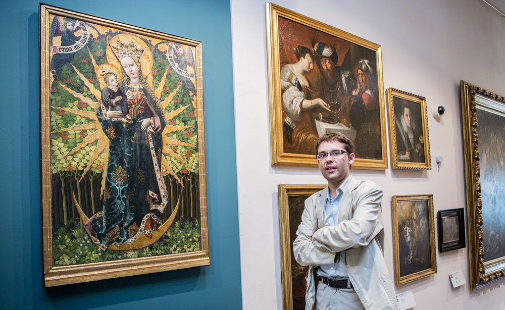 Mężczyzna w jasnej marynarce i okularach stoi z założonymi ramionami w galerii przy ścianie z obrazami; po lewej duży religijny obraz kobiety trzymającej dziecko na niebiesko-zielonym tle z złotą aureolą, po prawej kilka mniejszych, złoconych ramek na jasnej ścianie i widoczna kamera nad nimi.