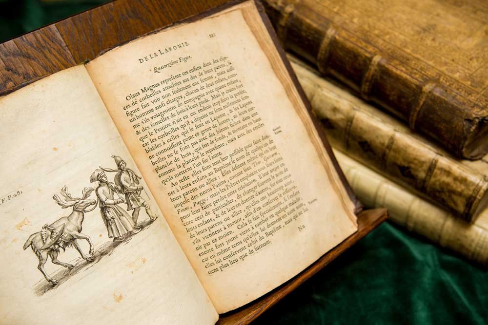 Otwarte stare wydanie: prawa strona z francuskim tekstem i tytułem "DE LA LAPONIE", lewa ze szkicowaną ilustracją dwóch postaci prowadzących renifera, w tle stos skórzanych opraw książek na zielonym materiale.