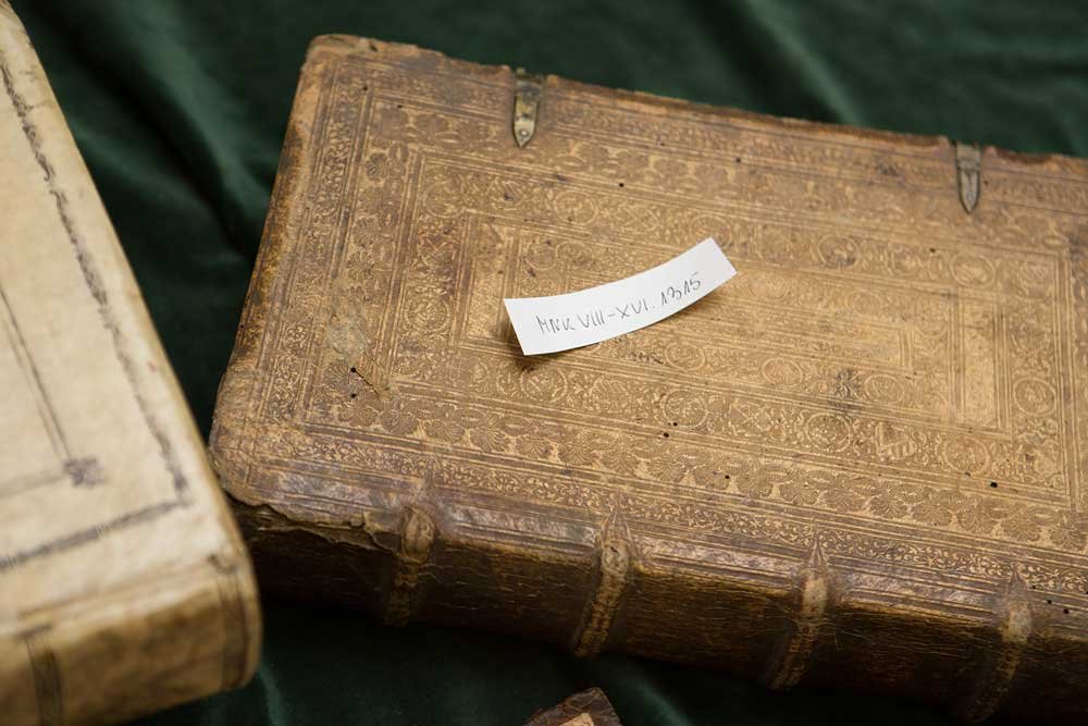 Duża ozdobna książka w brązowej skórzanej oprawie z tłoczonymi ornamentami i metalowymi okuciami leżąca na zielonym materiale, na okładce mała papierowa karteczka z odręcznym napisem, u dołu z lewej fragment innej jasnej księgi.