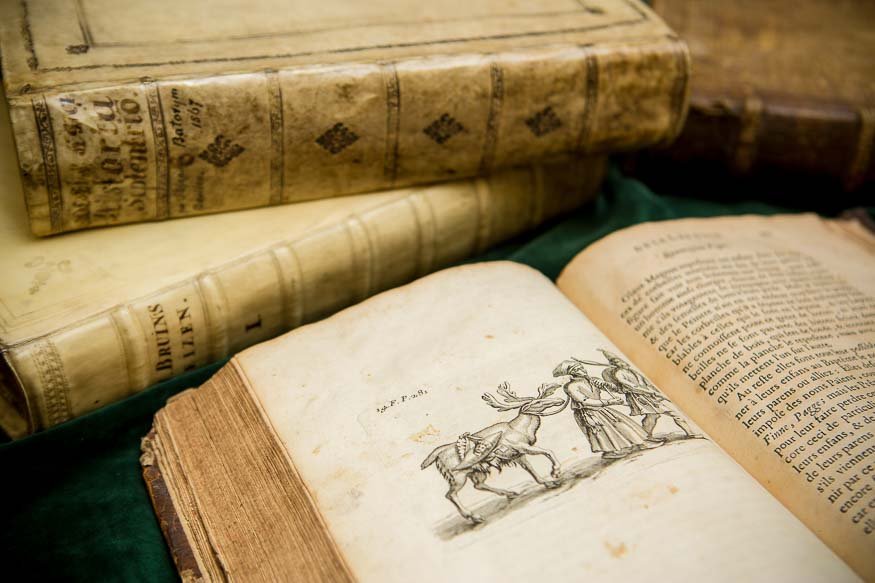Stare, oprawione książki ułożone na zielonym materiale, z jedną otwartą na ilustracji przedstawiającej jelenia prowadzonego przez dwie postacie oraz zadrukowaną stroną z tekstem; widoczne pożółkłe, postrzępione karty i ozdobne grzbiety.
