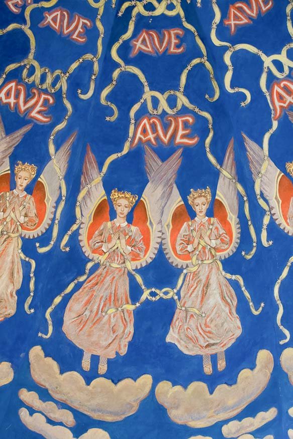 Powtarzający się ornament przedstawiający anioły w koronach w różowych szatach i czerwono-białych skrzydłach z złożonymi rękami, otoczone splecionymi złotymi wstążkami i czerwonym napisem "AVE" na intensywnie niebieskim tle nad chmurami.