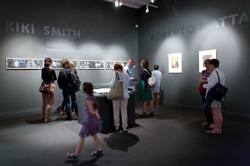 Wnętrze ciemnej galerii z kilkoma osobami oglądającymi prace na ścianach — po lewej pozioma seria oprawionych grafik pod napisem "KIKI SMITH", po prawej dwie oprawione prace pod napisem "ROBERTO MATTA", w środku przeszklona gablotka, a na pierwszym planie dziewczynka w fioletowej sukience idąca przez salę.