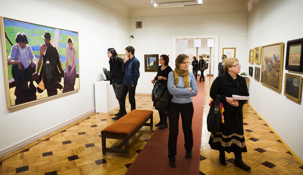 Wnętrze galerii sztuki z białymi ścianami i parkietem, zwiedzający oglądający obrazy — po lewej duży kolorowy obraz z trzema postaciami, przed nim dwóch mężczyzn stojących przy białym postumencie i pomarańczowa ławka, w środku kobieta w szarym swetrze i żółtym szaliku ze skrzyżowanymi ramionami, po prawej kobieta w czarnym ubraniu trzymająca dokumenty i na dalszym planie kolejni odwiedzający oraz obrazy w ramkach.