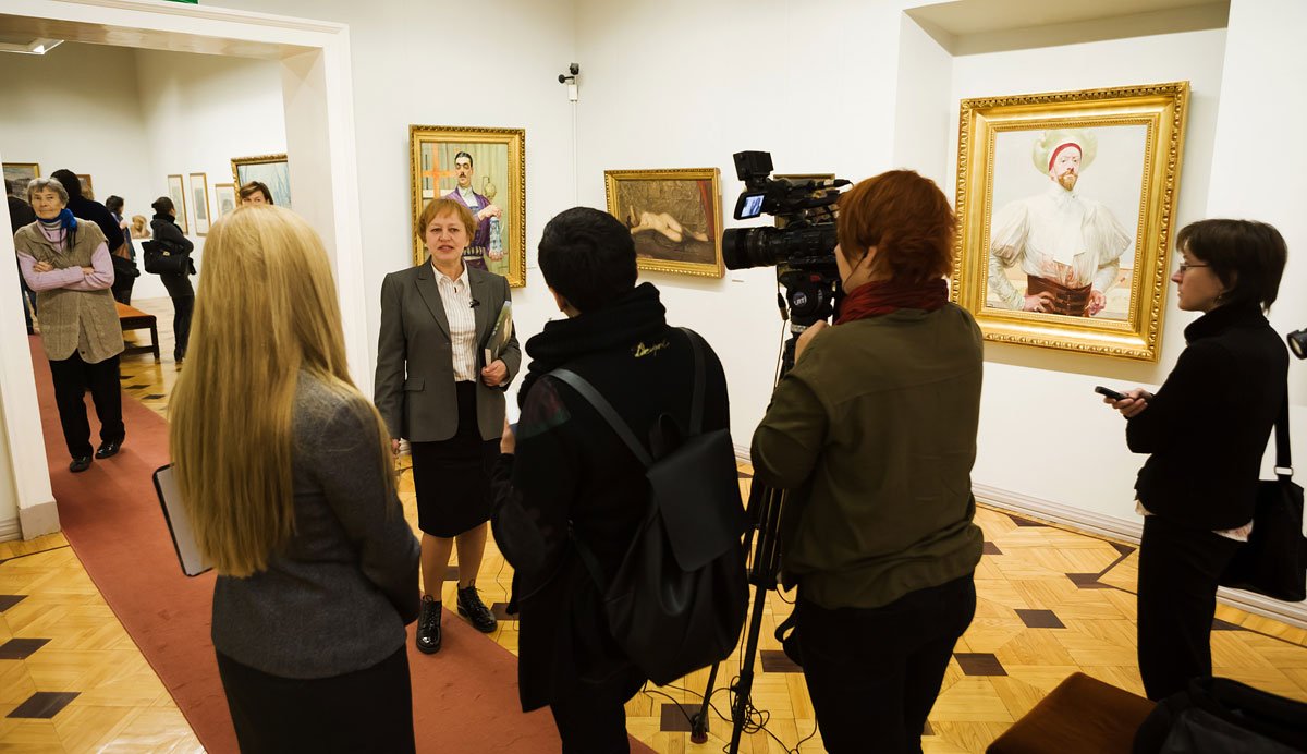 Wnętrze galerii sztuki z mozaikową drewnianą podłogą i czerwonym dywanem, gdzie kobieta w garniturze stoi przy ścianie z oprawionymi obrazami, przed nią operator z profesjonalną kamerą oraz kilka osób z notatnikami i telefonami, a w tle inni zwiedzający.