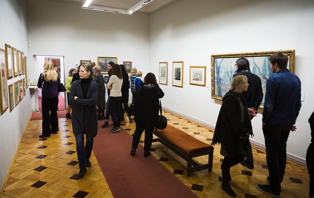 Wnętrze jasnej galerii z drewnianą podłogą i czerwonym dywanem, na ścianach oprawione obrazy i grafiki, w sali kilkanaście osób stojących przy obrazach oraz drewniana ławka pośrodku.