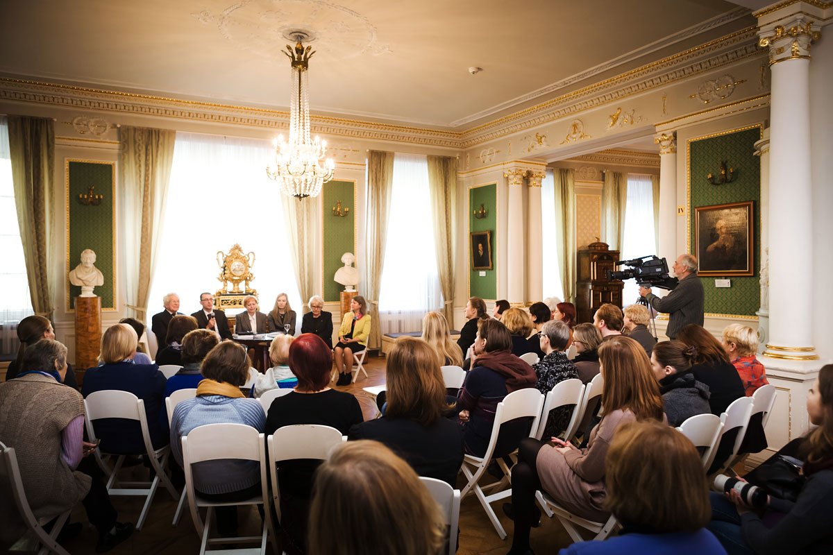 Duża grupa osób siedzących na białych składanych krzesłach ogląda panel kilku prelegentów przy stole w elegancko zdobionym salonie z kryształowym żyrandolem, wysokimi oknami i zielonymi boazeriami, marmurowymi popiersiami oraz operatorem kamery filmującym z prawej strony.