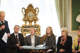 Grupa sześciu osób w eleganckim pomieszczeniu: po lewej stoi starszy mężczyzna trzymający mikrofon, przy stoliku siedzi mężczyzna i cztery kobiety, na okrągłym stoliku leżą dokumenty i szklanki z wodą, a w tle na środku stoi duży złocony ozdobny zegar na podstawie przed zasłonami.