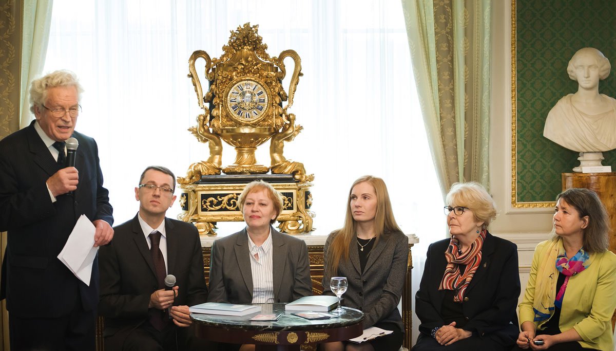 Sala reprezentacyjna z sześcioma osobami przy okrągłym stoliku: stojący po lewej starszy mężczyzna z mikrofonem i kartkami, obok siedzi mężczyzna w garniturze trzymający mikrofon, dalej trzy kobiety w formalnych strojach, na stole dokumenty i szklanka wody, a w tle duży złocony ozdobny zegar, grube zasłony i marmurowe popiersie.
