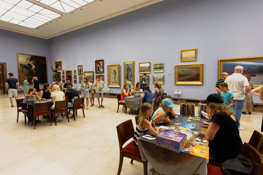 Duża sala galerii ze ścianą w jasnym fiolecie z wieloma obrazami w złotych ramach i dużym świetlikiem sufitowym, w której dorośli i dzieci siedzą przy stołach grając w gry planszowe na pierwszym planie, a inni zwiedzający stoją i spacerują przy eksponatach.
