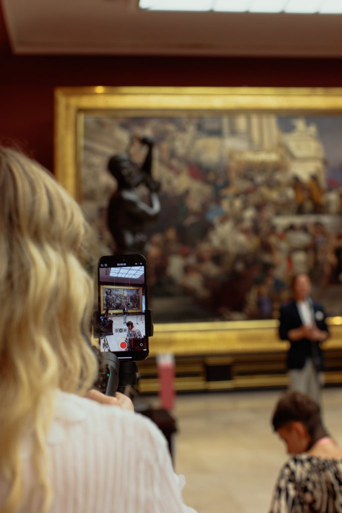 Osoba z blond włosami stoi tyłem i filmuje smartfonem zamocowanym na gimbalu duże, wielobarwne malowidło w złotej ramie w galerii; na ekranie telefonu widoczny jest podgląd nagrania z ciemną rzeźbą i kilkoma osobami, a oryginalny obraz i rozmyte sylwetki w tle są poza ostrością.