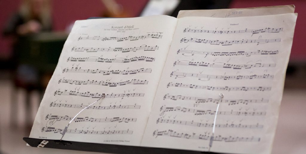 Otwarta partytura na pulpicie nutowym — dwie strony z pięcioliniami, nutami i odręcznymi znakami oraz widocznym tytułem Konzert d‑Moll u góry, przytrzymana metalowymi klipsami i uchwytami, na rozmytym tle zarysowane postaci i instrument.