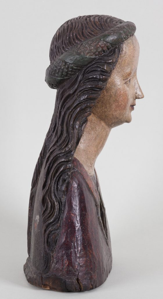 Drewniana rzeźba popiersia kobiety ukazana w profilu zwróconym w prawo, z długimi falistymi włosami oplecionymi warkoczem na czubku głowy, pomalowaną, częściowo startego beżowego koloru twarzą z zaznaczonymi różowymi policzkami i ustami, długą szyją, ciemnoczerwoną szatą oraz widocznymi pęknięciami i śladami zużycia na podstawie i powierzchni.