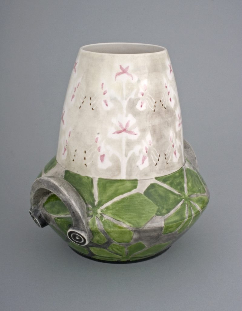 Ceramiczny dzban z wąską białą szyjką ozdobioną drobnymi różowymi kwiatowymi motywami i kropkowanymi detalami, szerokim korpusem w zielone płatkowate wzory oraz szarą spiralną rączką, na jednolitym szarym tle.