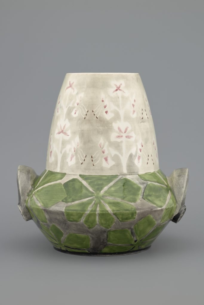 Fragment ceramicznego wazonu o zaokrąglonym korpusie i zwężonej szyjce z dwiema uszami; dolna część pokryta zielonymi geometrycznymi wzorami przypominającymi liście, górna w jasnoszarym kolorze z drobnymi różowymi motywami kwiatowymi, na jednolitym szarym tle.