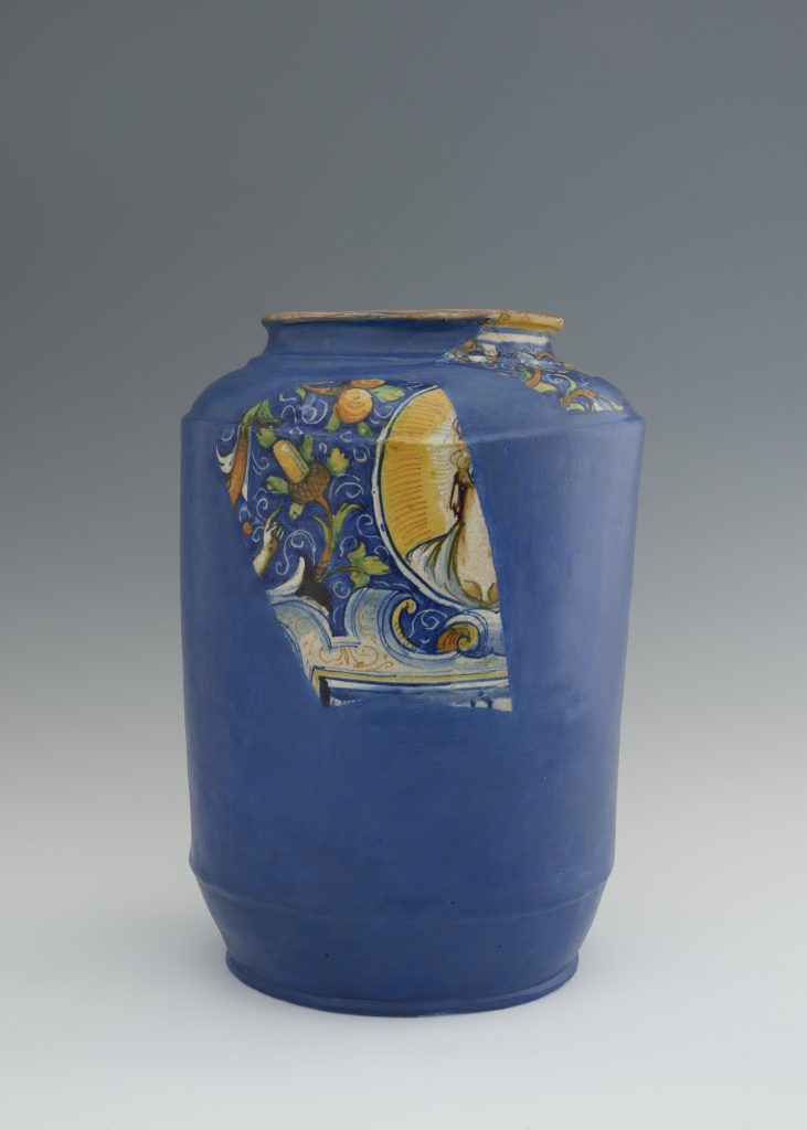 Wysoki cylindryczny ceramiczny wazon w intensywnym kobaltowym niebieskim na gładkim szarym tle, z ubytkami przy szyjce i widocznym fragmentem wielobarwnej dekoracji roślinno‑figuralnej w żółtych, zielonych i pomarańczowych tonach.