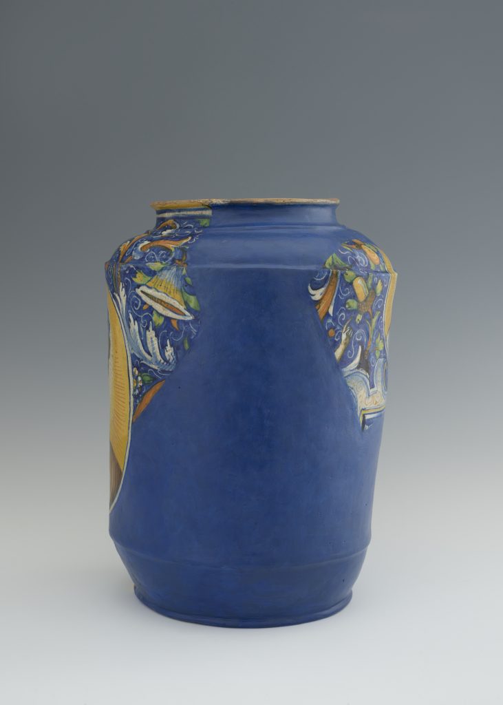 Wysoki matowy niebieski ceramiczny wazon o szerokim korpusie, z ozdobnymi kolorowymi motywami roślinnymi w polach przy szyjce i bokach (żółć, zieleń, biel) oraz lekko przetartą krawędzią wylewu odsłaniającą surową glinę, na neutralnym szarym tle.