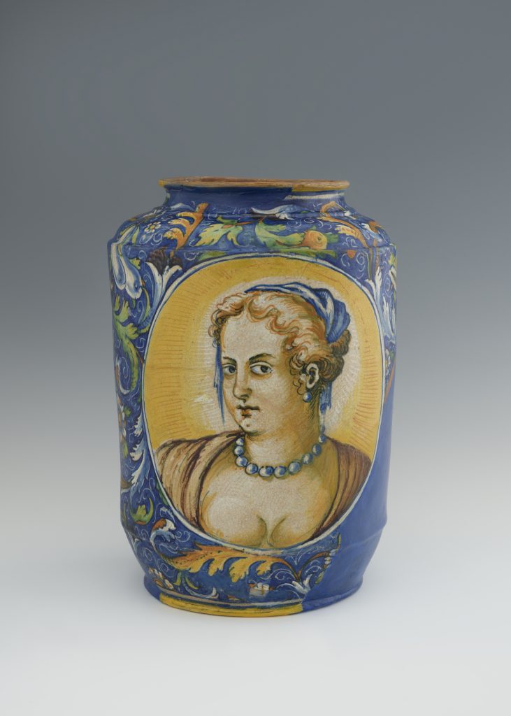 Fragment wysokiego ceramicznego naczynia w głębokim kobaltowym kolorze z bogatą roślinną ornamentyką w zieleni i złocie oraz dużym owalnym medalionem przedstawiającym popiersie kobiety z kręconymi włosami, chustką na głowie i naszyjnikiem z kulistych korali na żółtym tle, ze spękaniami szkliwa i drobnymi ubytkami przy rancie.