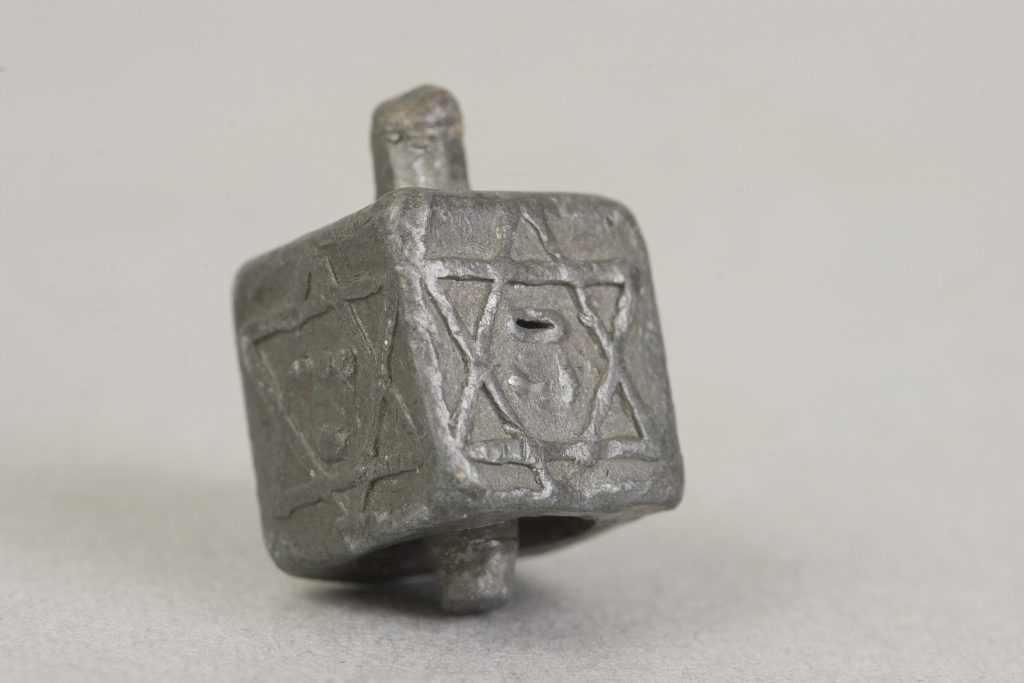 Zbliżenie szarego metalowego sześciennego przedmiotu z wyrytymi geometrycznymi znakami na bokach, małym otworem i krótkim wystającym trzpieniem, na jasnym neutralnym tle.