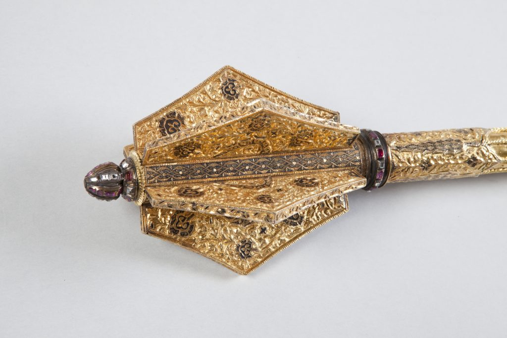 Fragment ozdobnego pozłacanego przedmiotu z wielofasetową, sześciokątną głowicą o wypukłych płatach zdobionych misterną roślinną repusowaną ornamentyką i czarnymi emaliowanymi motywami, z metalowym guzikiem wysadzanym fioletowymi kamieniami, pierścieniem z czerwonych kamieni oraz rytowanym trzonkiem, na jasnoszarym tle.