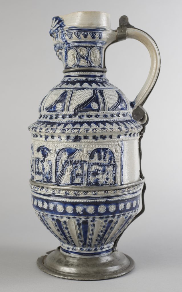 Ceramiczny, wysoki dzban z niebiesko-białym szkliwem i metalowym (cynowym) uchwytem oraz zawiasem przy szyjce, bogato zdobiony reliefowymi pasami: miniaturowe scenki w arkadach i medaliony na górze, rzędy drobnych rozet i fryzów oraz żłobione pionowe żebra na dolnej części naczynia.