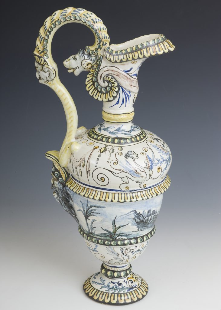 Ozdobny ceramiczny dzban w stylu majoliki z wysoką szyjką i nóżką, z ozdobnym wygiętym uchwytem zdobionym dwiema twarzami i zwierzęcą głową przy wylewie, bogato malowany ornamentami, pasami perełek i motywami roślinnymi, pejzażami oraz figurami w odcieniach niebieskiego, żółtego, zielonego i czarnego.