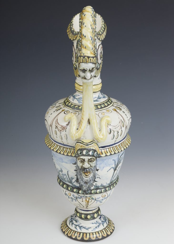Ozdobny ceramiczny wazon z pokrywką na jednolitym szarym tle, biało‑żółto‑niebieskie szkliwienie, pokrywka zakończona reliefową twarzą z wydłużonym, skręconym elementem i stylizowanymi uszami, na korpusie druga wypukła brodata twarz, liczne motywy roślinne oraz pasy z zielono‑żółtymi punktami i złoto‑żółtymi akcentami.