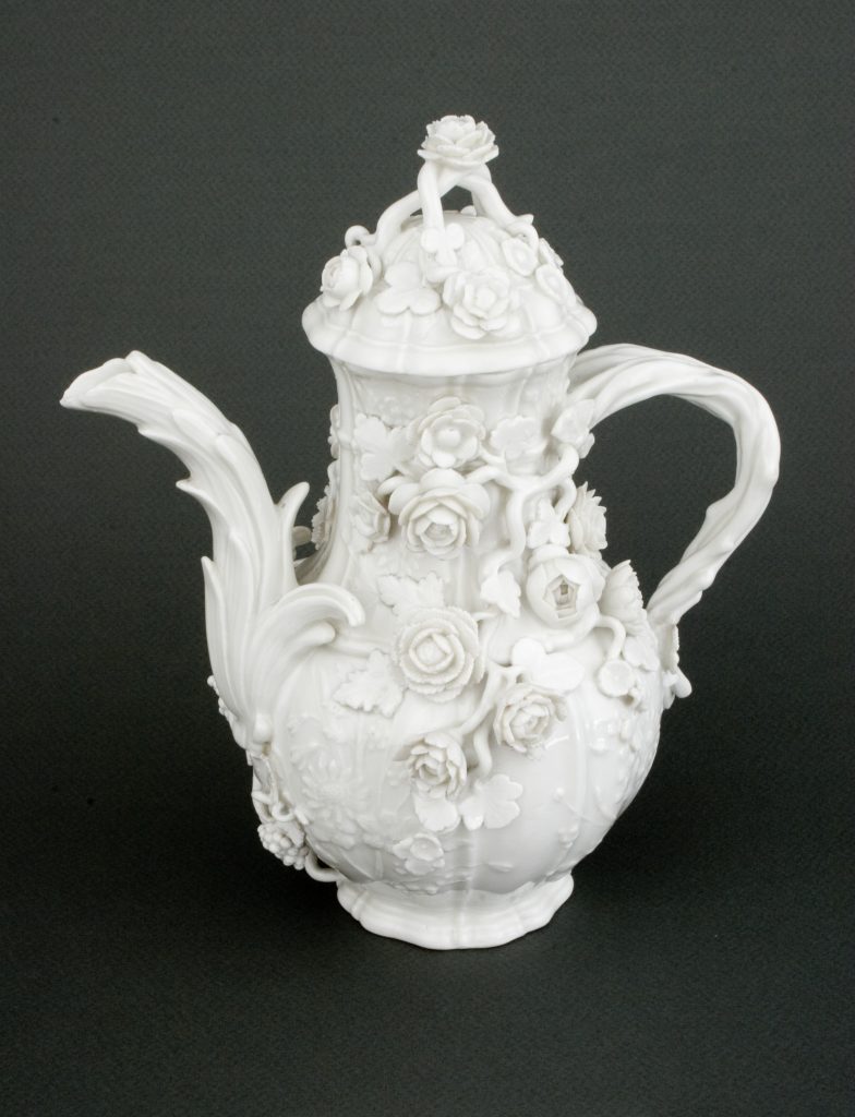 Biały porcelanowy imbryk bogato zdobiony reliefowymi różami i liśćmi, z pokrywką zakończoną różą, ozdobnym zakrzywionym dzióbkiem i uchwytem, na ciemnoszarym tle.