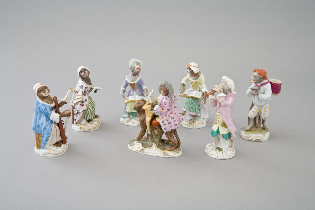 Siedem drobnych porcelanowych figurek przedstawiających małpy w strojach z epoki, ustawionych w półokręgu na jednolitym szarym tle, niektóre trzymają instrumenty (harfa, bęben, flet) lub śpiewniki, każda na zdobionej, częściowo złoconej podstawie.