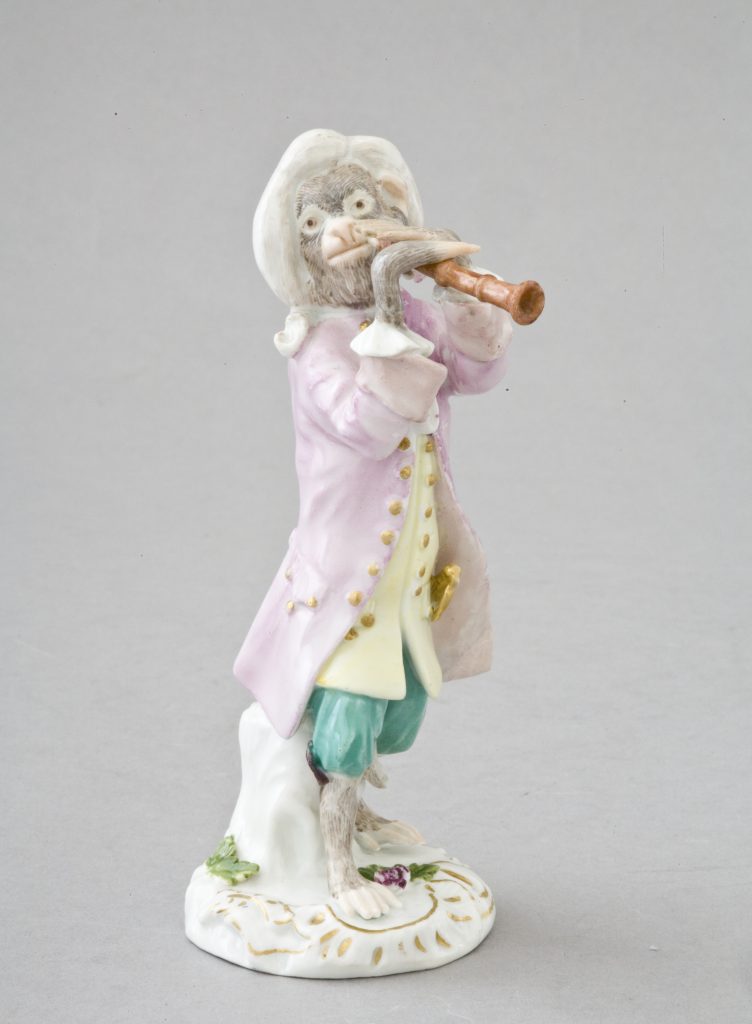 Porcelanowa figurka antropomorficznej małpy w białej peruce i pastelowym stroju — różowy płaszcz, żółta kamizelka i zielone spodnie — stojąca na ozdobnej białej podstawce ze złotymi zdobieniami i kwiatem, trzymająca przy ustach zakrzywiony drewniany instrument dęty, z widocznym futrzastym ciałem i gołymi stopami.