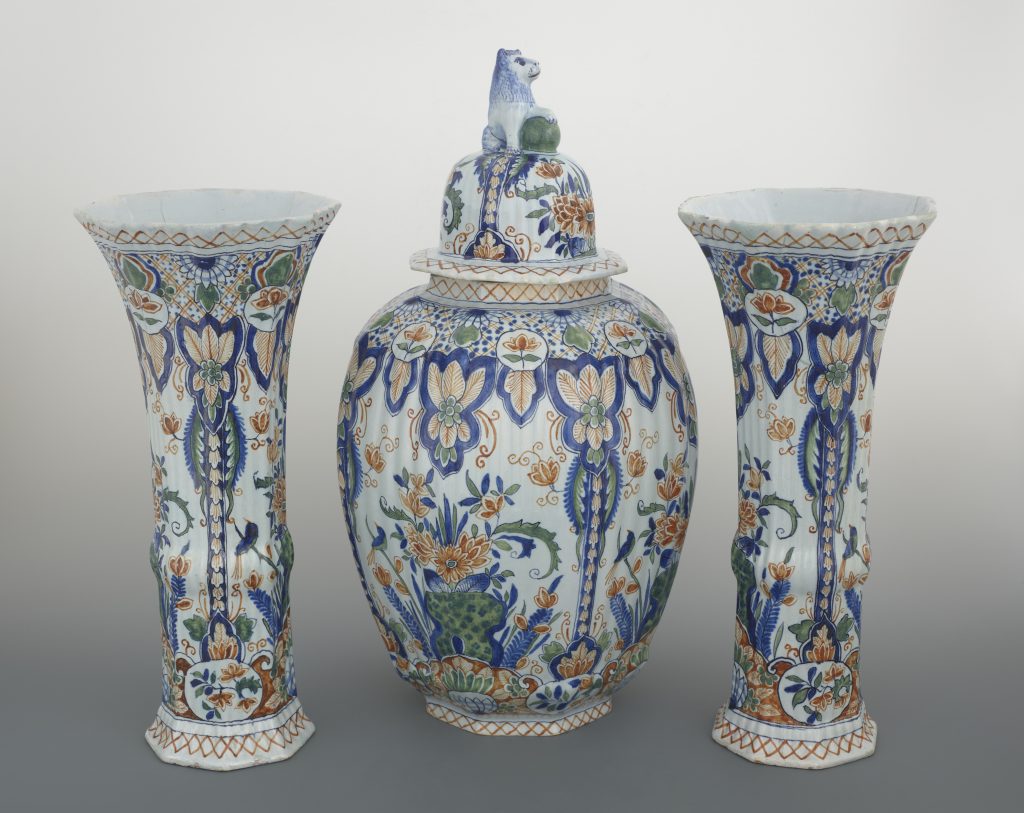 Trzy ceramiczne naczynia na jasnym tle: centralny przykryty wazon z bogatym roślinnym wzorem w odcieniach niebieskiego, zielonego i pomarańczowego oraz pokrywą zwieńczoną małą figurką zwierzęcia trzymającego kulę, po obu stronach dwie wysokie, ku górze rozszerzane wazy z tym samym motywem dekoracyjnym.