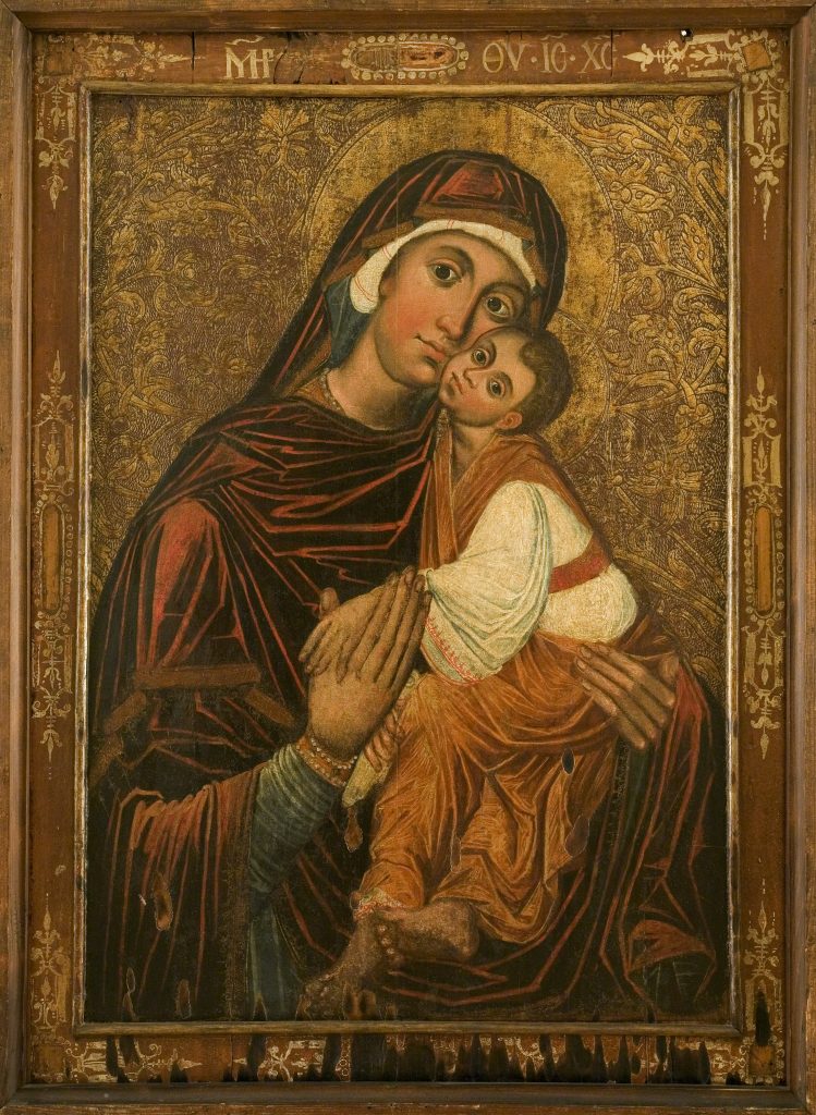 Malarska ikona w ozdobnej drewnianej ramie ukazująca pochyloną kobietę w ciemnoczerwonym płaszczu trzymającą blisko siebie małe dziecko w białej tunice i pomarańczowym okryciu na złocistym, ornamentalnym tle z aureolami.