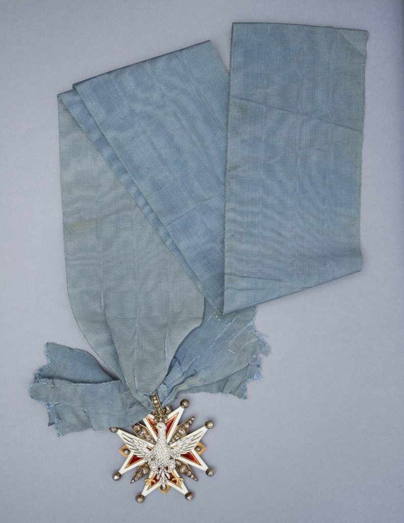 Jasnoniebieska, drobno prążkowana wstęga z załamaniami i postrzępionymi końcami leżąca na jasnoszarym tle, u dołu przywiązany gwiaździsty medal z białą emaliowaną figurą ptaka o rozpostartych skrzydłach na czerwono-białej gwieździe oraz metalowymi kulkami i ozdobnymi kamieniami.