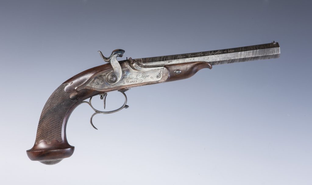 Zbliżenie na zabytkowy pistolet z ciemnym, żłobionym drewnianym chwytem, metalową okładziną z wygrawerowanymi dekoracjami, widocznym młoteczkiem i podwójnym spustem oraz długą ośmiokątną lufą z grawerowanym napisem na jasnym neutralnym tle.