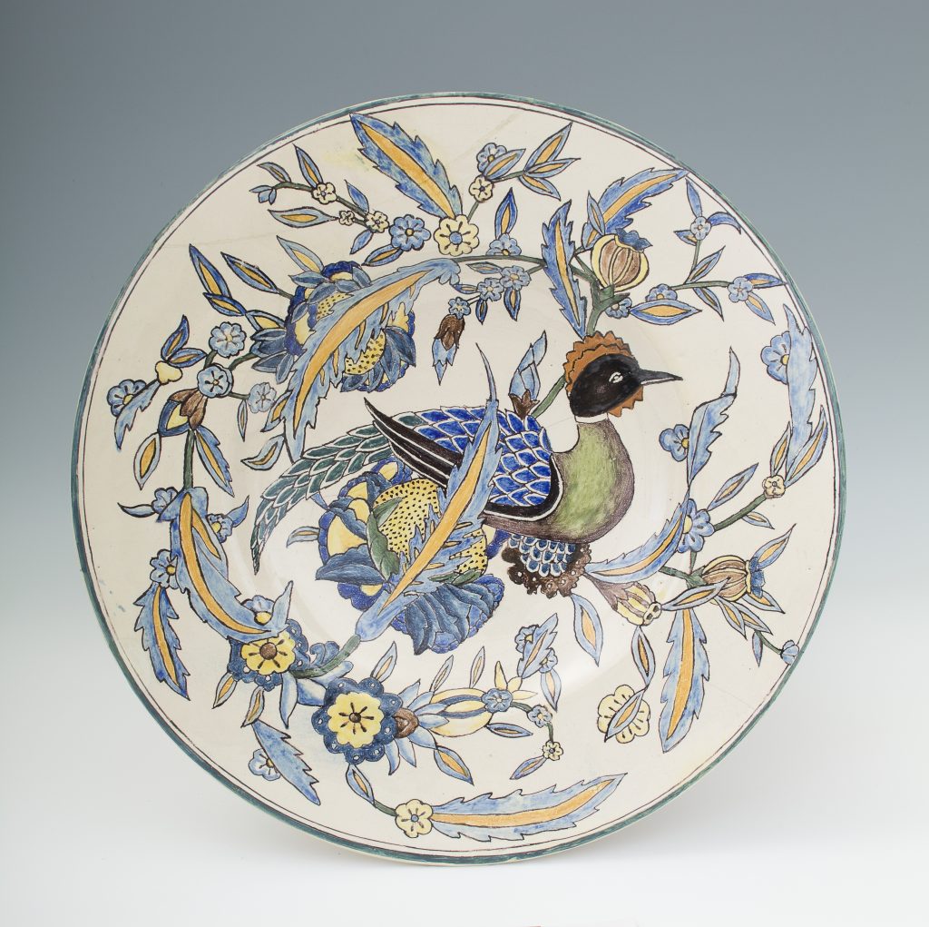 Okrągły ceramiczny talerz z malowanym, stylizowanym ptakiem i rozgałęzionym ornamentem kwiatowo‑liściastym w odcieniach niebieskiego, żółtego, zielonego i czarnego na jasnym tle z cienką zieloną obwódką brzegu.