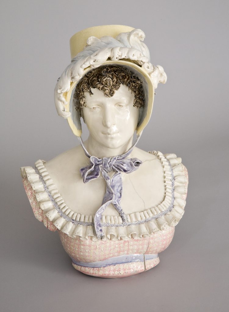 Porcelanowe popiersie kobiety ukazujące głowę i tors, z kręconymi ciemnymi włosami pod żółtym kapeluszem ozdobionym białymi falistymi detalami, z fioletową wstążką zawiązaną pod szyją oraz szerokim falbaniastym dekoltem i różowo-wzorowanym gorsetem.