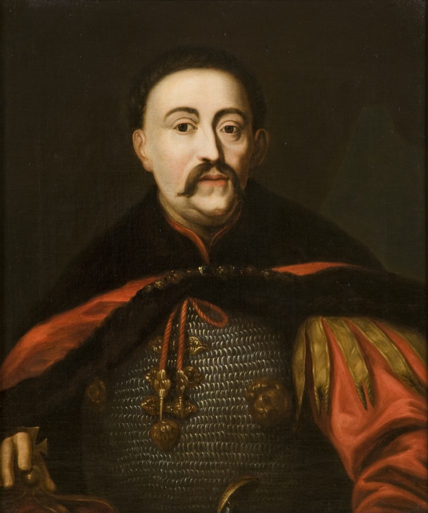 Popiersiowy portret mężczyzny na ciemnym tle ukazujący zbliżenie twarzy i tułowia w kolczudze z ozdobnymi złotymi okuciami i czerwonym sznurem, przepasanego czerwonym płaszczem z pasami w złotym odcieniu, z krótkimi ciemnymi włosami, wąsami i niewielką brodą.