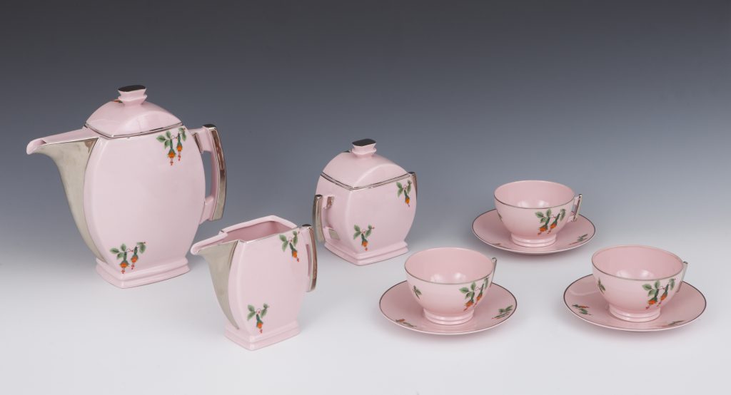 Różowy porcelanowy serwis herbaciany rozłożony na jasnoszarym tle, składający się z dzbanka, mlecznika, cukiernicy oraz trzech filiżanek z podstawkami, wszystkie o geometrycznych kształtach z metalicznymi srebrnymi okuciami i drobnym motywem zielonych liści z pomarańczowymi kwiatuszkami.