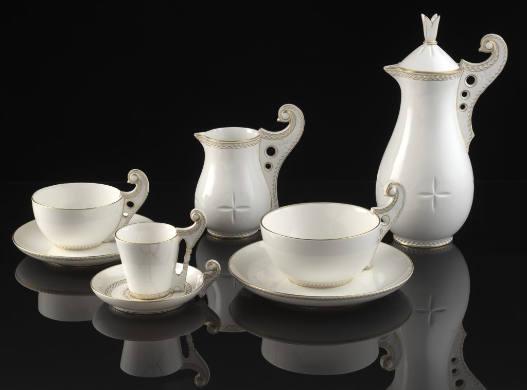 Biały porcelanowy serwis kawowy z filiżankami i spodkami, mlecznikiem, cukiernicą i wysokim dzbankiem z pokrywką ustawiony na czarnej błyszczącej powierzchni; widoczne zakręcone uchwyty z otworami, złote obwódki i zygzakowate zdobienia oraz tłoczone gwiazdkowe motywy, z delikatnymi odbiciami w lustrze.