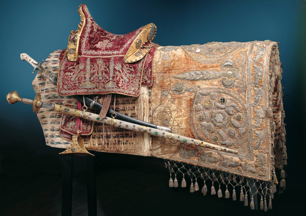 Detaliczne zdjęcie ozdobnego, historycznego siodła na stojaku: czerwone aksamitne siodło z pozłacanymi okuciami i srebrnym haftem, obok bogato haftowana złoto‑beżowa kapa z metalowymi aplikacjami i frędzlami oraz poprzeczne, zdobione miecze i pochwy z inkrustacjami.