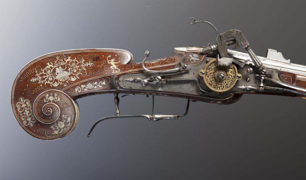 Fragment bogato zdobionego historycznego pistoletu: spiralna, ciemnobrązowa kolba z białymi inkrustacjami i ornamentami roślinnymi oraz figurkami, obok skomplikowany metalowy mechanizm z grawerowanym, mosiężnym kołem, stalowymi sprężynami i zamkiem.