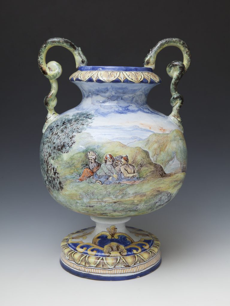 Ceramiczny ozdobny wazon na nodze o kulistym korpusie, z dwoma zielonkawymi spiralnymi uchwytami, niebieskim rantem zdobionym żółtym motywem liści, malowanym krajobrazem z grupą postaci w turbanach na boku oraz podstawą udekorowaną błękitno-żółtymi ornamentami.