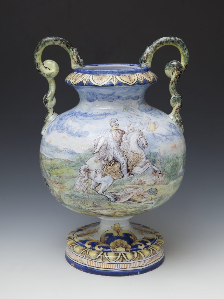 Duży zdobiony ceramiczny wazon na nodze z parą zielonych, spiralnie zakręconych uchwytów przypominających węże, niebieskim rantem ozdobionym beżowo-złotymi liśćmi, malowaną sceną rycerza na koniu na tle chmur i pejzażu oraz dekorowaną niebiesko-żółtą podstawą.