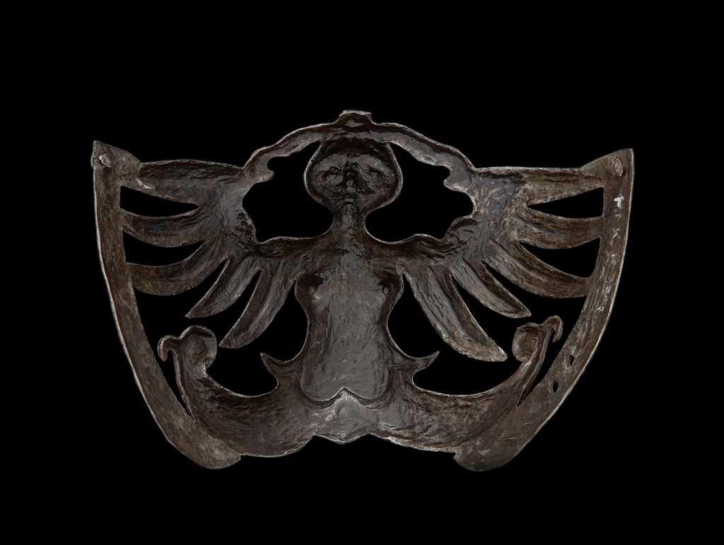Zbliżenie górnej części ciemnego, patynowanego metalowego ornamentu przedstawiającego uproszczoną sylwetkę postaci z wyciętymi, skrzydłopodobnymi płatami i chropowatą fakturą na czarnym tle.