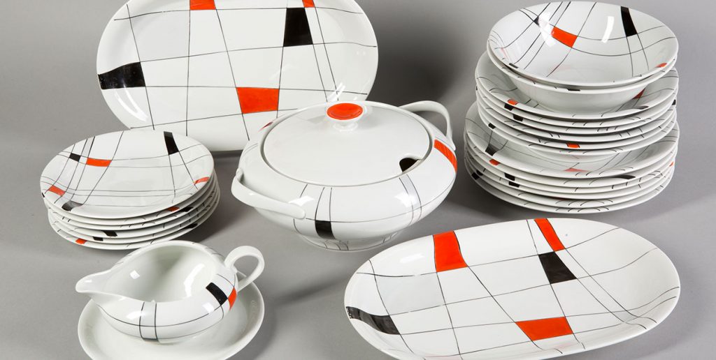 Ułożony na szarym tle komplet białej błyszczącej porcelany — stosy płaskich i głębokich talerzy, dwa owalne półmiski, waza z pokrywką i sosjerka na spodku — ozdobiony cienkimi czarnymi liniami tworzącymi siatkę oraz pojedynczymi pomarańczowymi i czarnymi prostokątnymi akcentami.