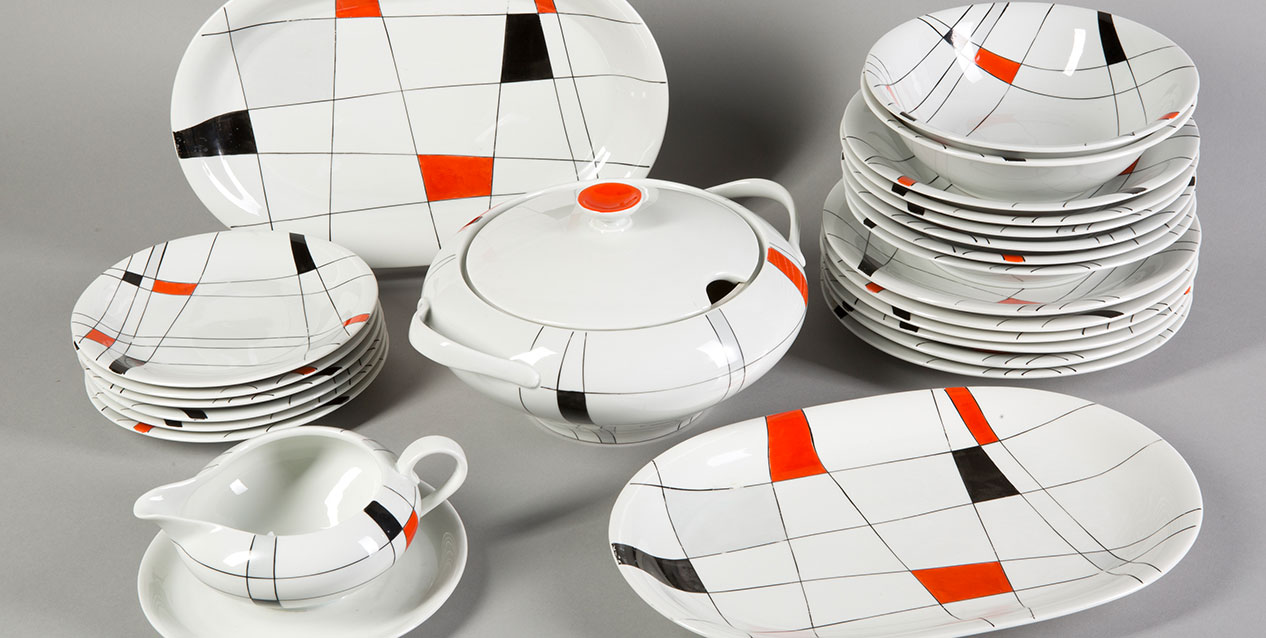 Ułożony na szarym tle komplet białej błyszczącej porcelany — stosy płaskich i głębokich talerzy, dwa owalne półmiski, waza z pokrywką i sosjerka na spodku — ozdobiony cienkimi czarnymi liniami tworzącymi siatkę oraz pojedynczymi pomarańczowymi i czarnymi prostokątnymi akcentami.