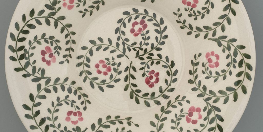 Fragment białego ceramicznego talerza zdobionego powtarzającym się wzorem zielonych, spiralnie wijących się gałązek z liśćmi i drobnymi różowo-czerwonymi kwiatkami na kremowym tle.