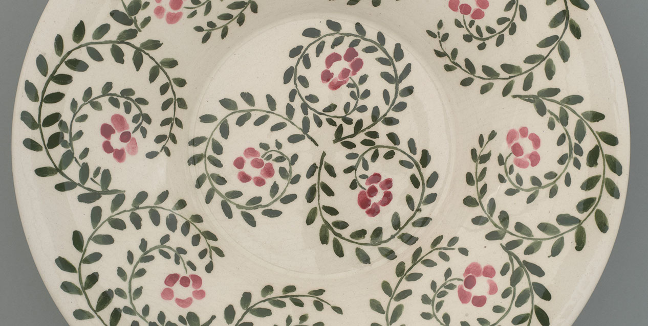 Fragment białego ceramicznego talerza zdobionego powtarzającym się wzorem zielonych, spiralnie wijących się gałązek z liśćmi i drobnymi różowo-czerwonymi kwiatkami na kremowym tle.